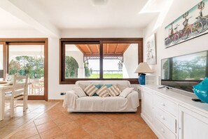Un salon avec un canapé, un téléviseur et une vue sur un jardin, visible à travers une grande fenêtre.