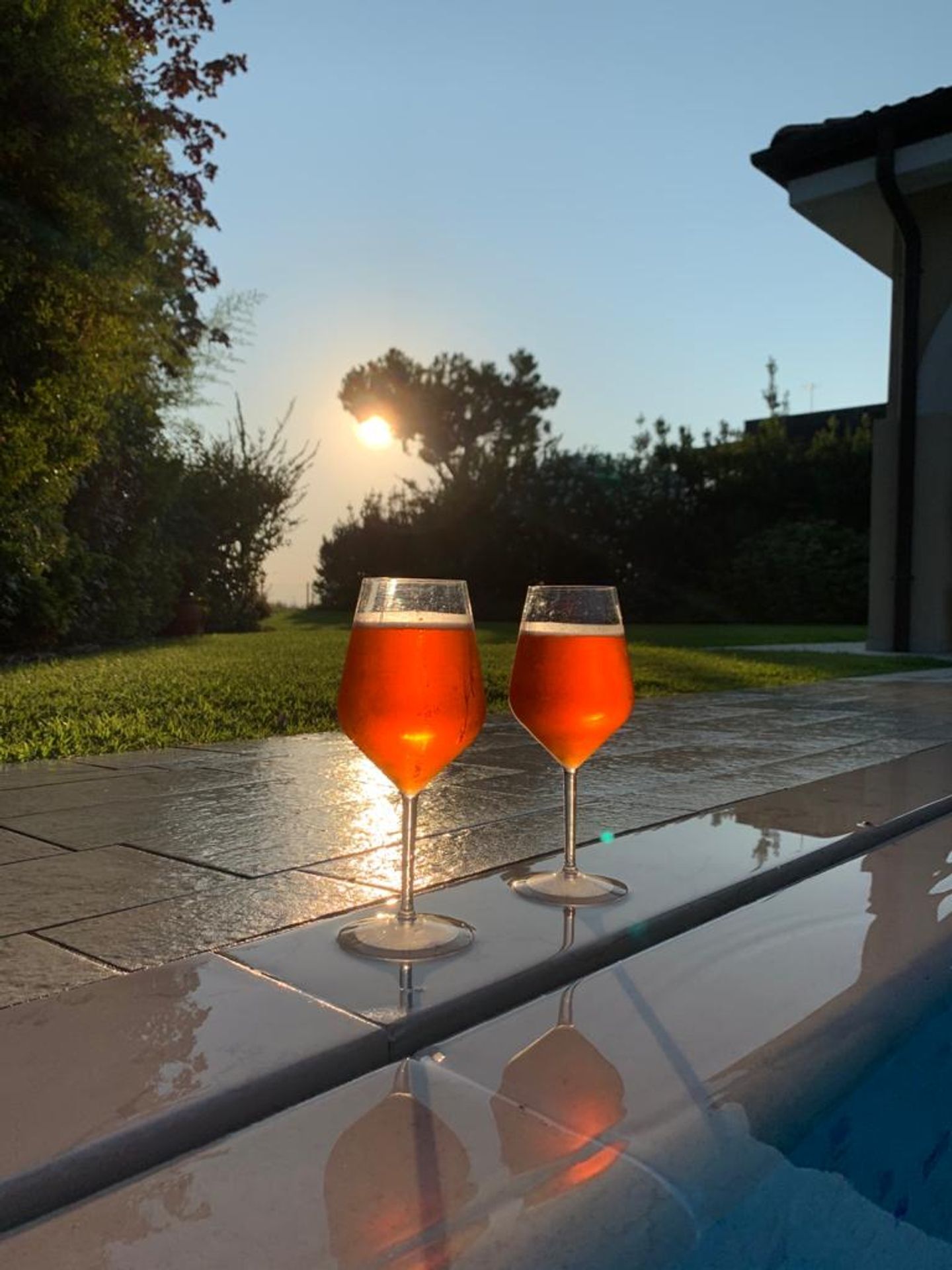 Two glasses of amber liquid on a reflective surface, likely a poolside area, with a sunset in the background.