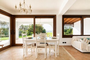 A dining area with a table set for four, a chandelier, and a view of greenery and a body of water through large sliding glass doors.