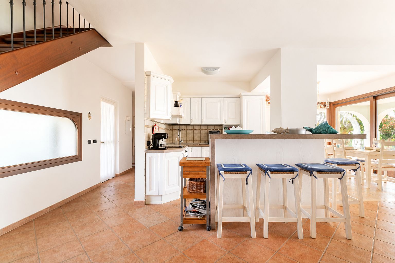 A kitchen with white cabinets, a wooden staircase, and a dining area with bar stools.