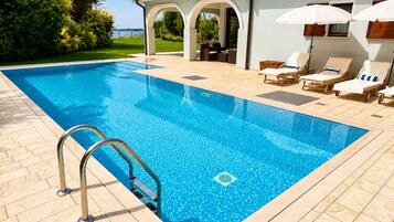 A swimming pool with a clear blue water, surrounded by a tiled deck, and a lounge area with chairs and umbrellas.