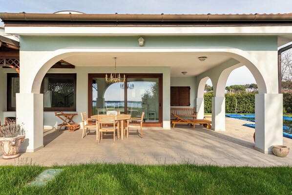 A covered patio area with a table and chairs, a hanging light fixture, and a view of the sea through large windows.