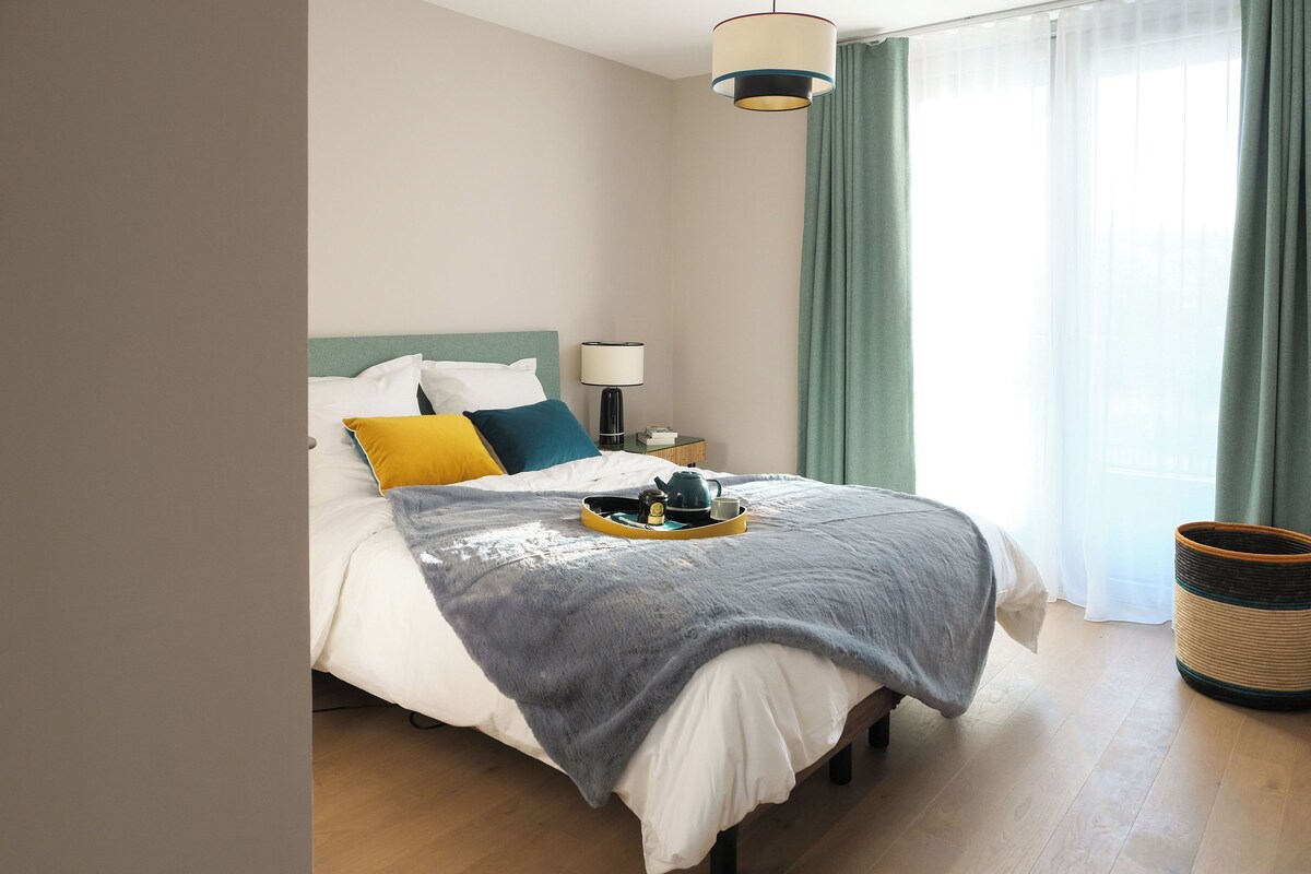 Un lit bien fait, avec des oreillers, une table de chevet sur laquelle se trouve une lampe, et un plateau avec une théière et une tasse.