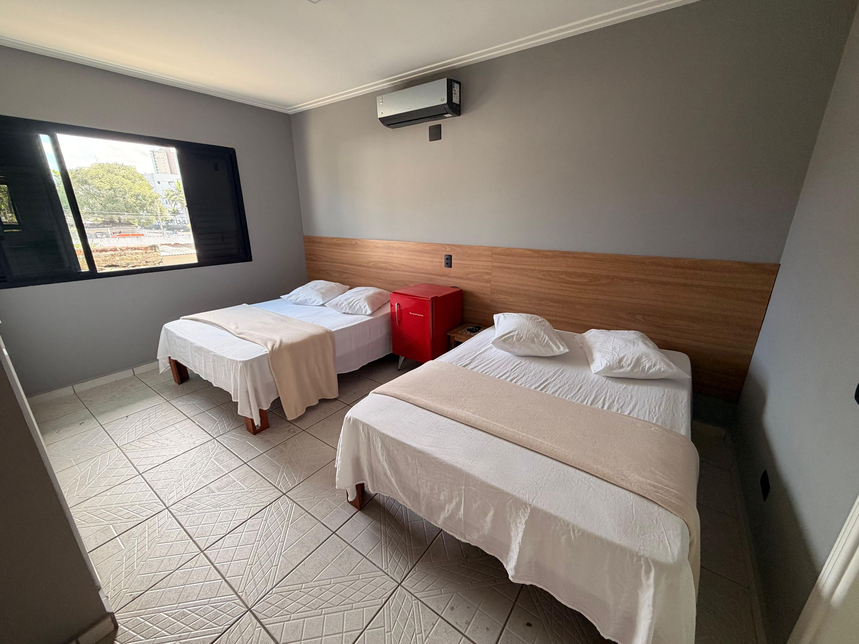 Quarto de hotel com duas camas, um minibar vermelho e janela com vista para edifícios.