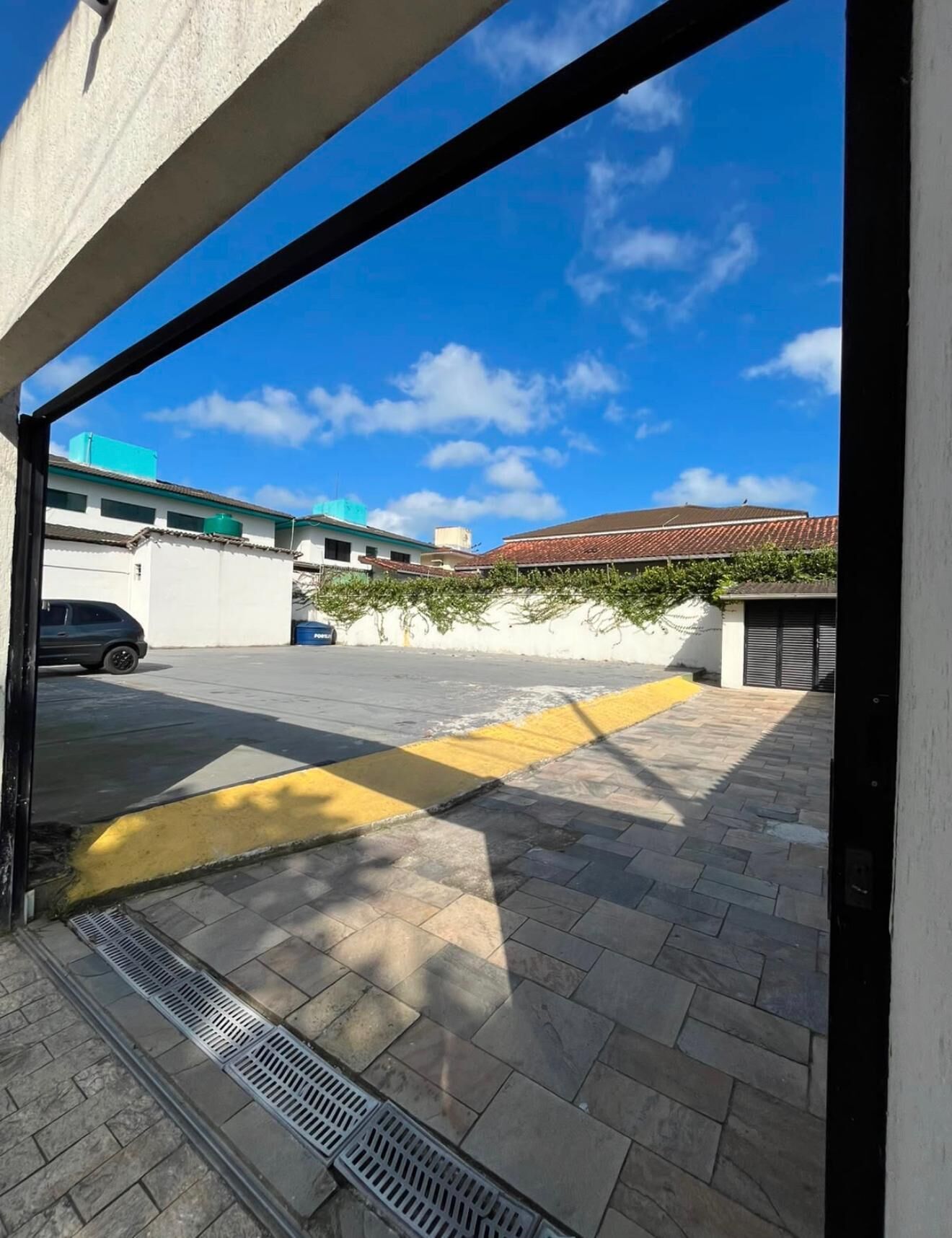 Un espacio exterior pavimentado con un camino de baldosas amarillas táctiles, una rejilla de drenaje y un edificio con techo de tejas rojas al fondo.