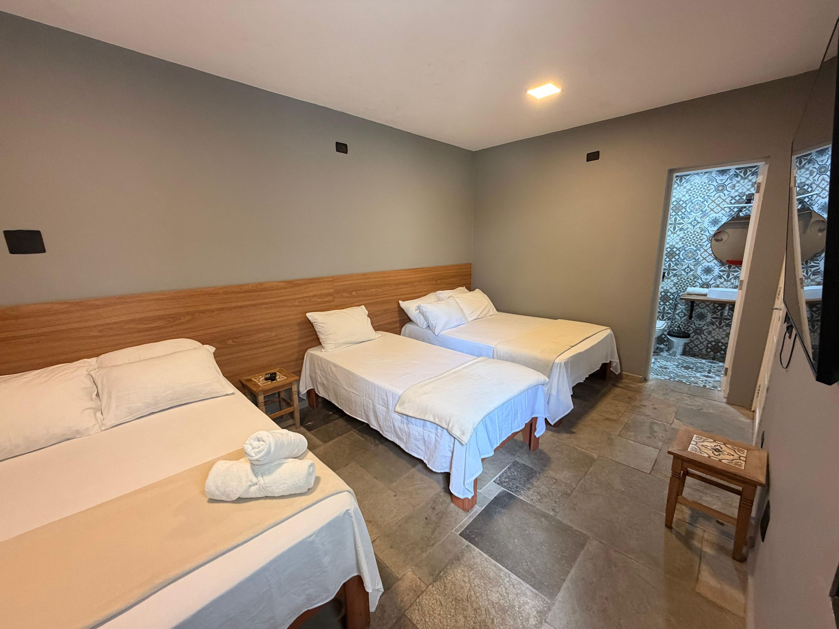 Ett hotellrum med tre sängar, en träpanel som huvudgavel, en liten träpall och ett badrum som syns genom en glasdörr.