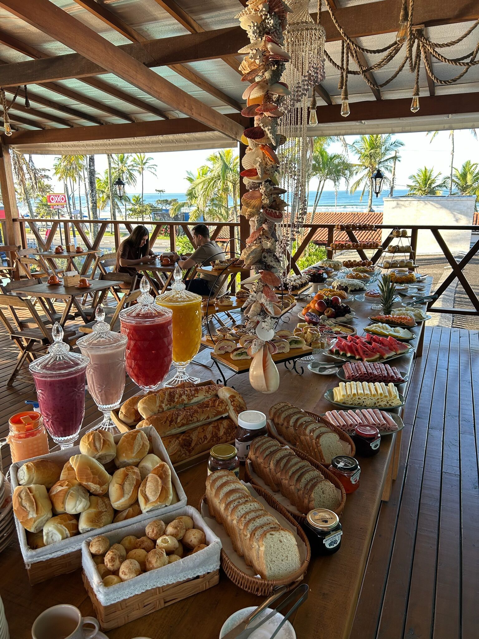 A buffet table with various food items and drinks, located under a wooden structure with a view of the beach and palm trees.