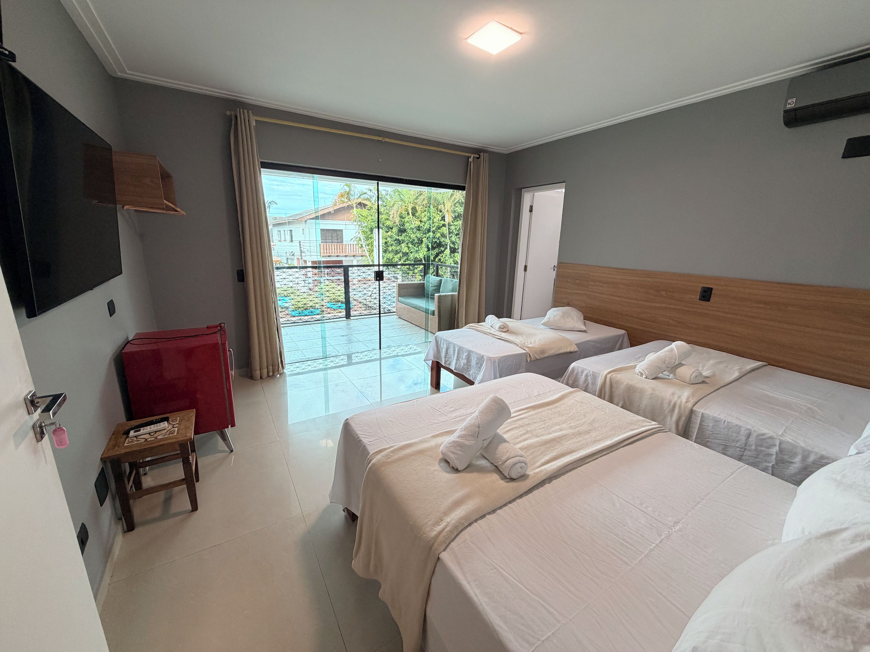 A hotel room with two beds, a red chair, a television, and a balcony with a view of buildings and greenery.
