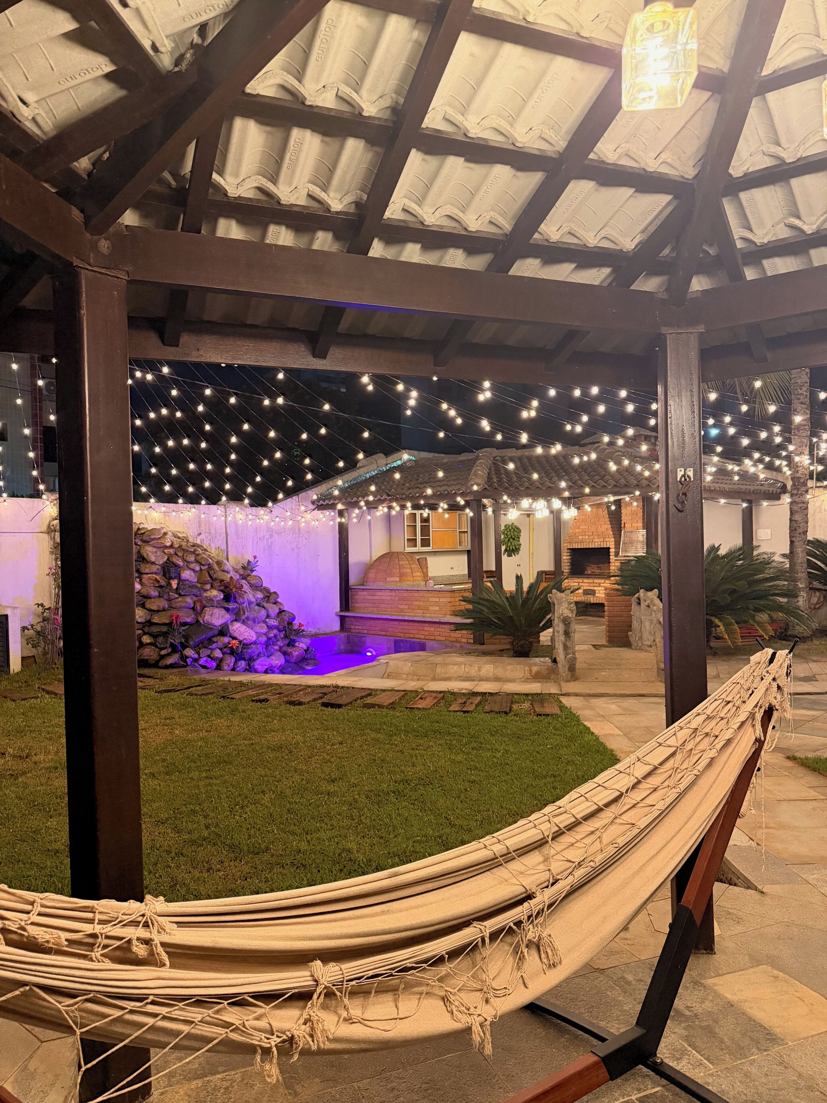Un hamac installé sous une zone abritée, agrémentée de guirlandes lumineuses, un four en briques et un petit jardin avec un élément décoratif aquatique.