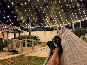 A hammock under string lights with a brick wall and palm trees in the background. - Santa Rita Pousada (Guarujá)