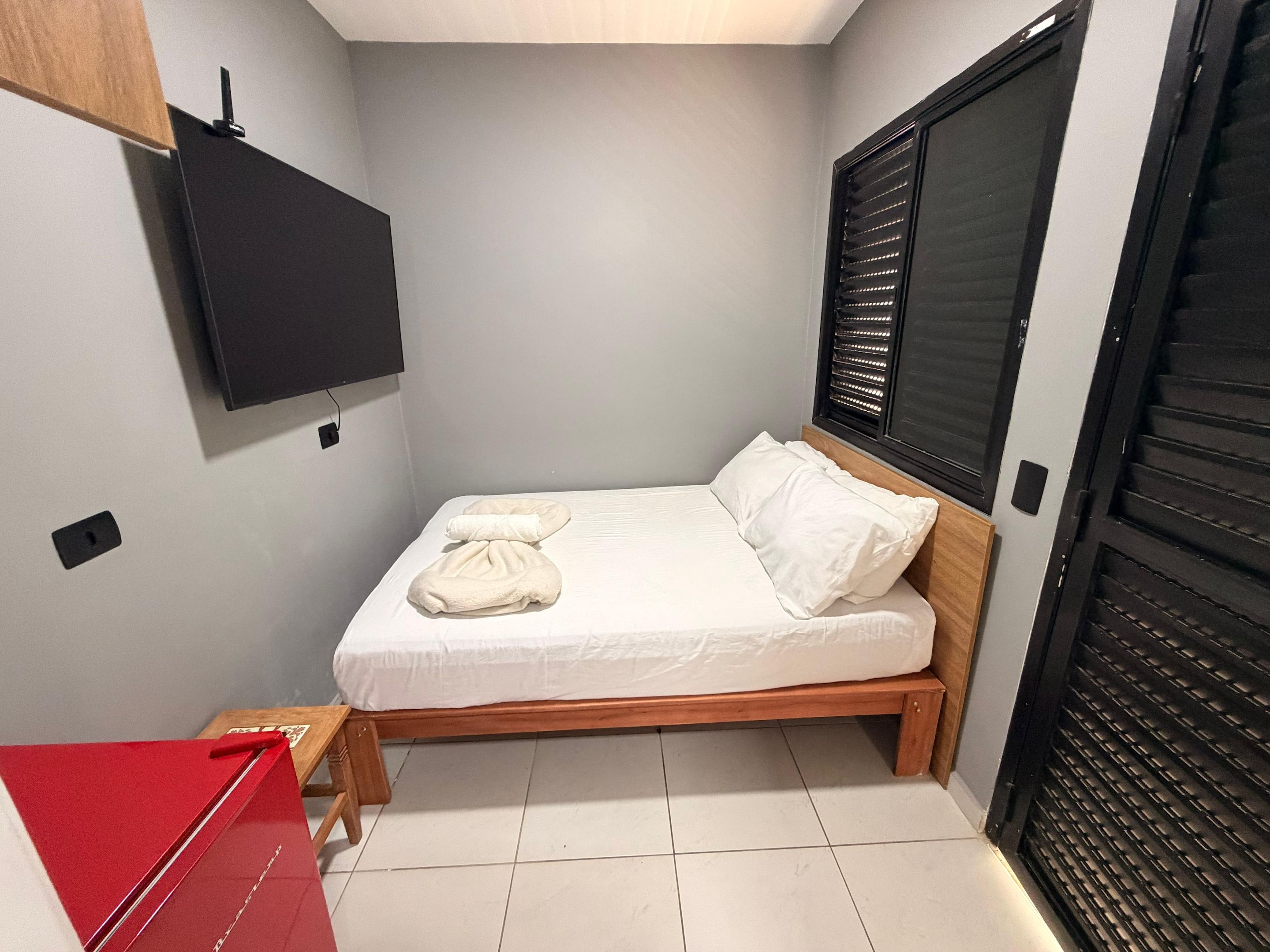 Un dormitorio pequeño y ordenado con una cama individual, cabecero de madera, una ventana con persianas y una carpeta roja sobre una mesita.