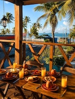Une table en bois, dressée avec des aliments pour le petit-déjeuner et du jus d’orange, donnant sur une plage bordée de palmiers et sur l’océan.