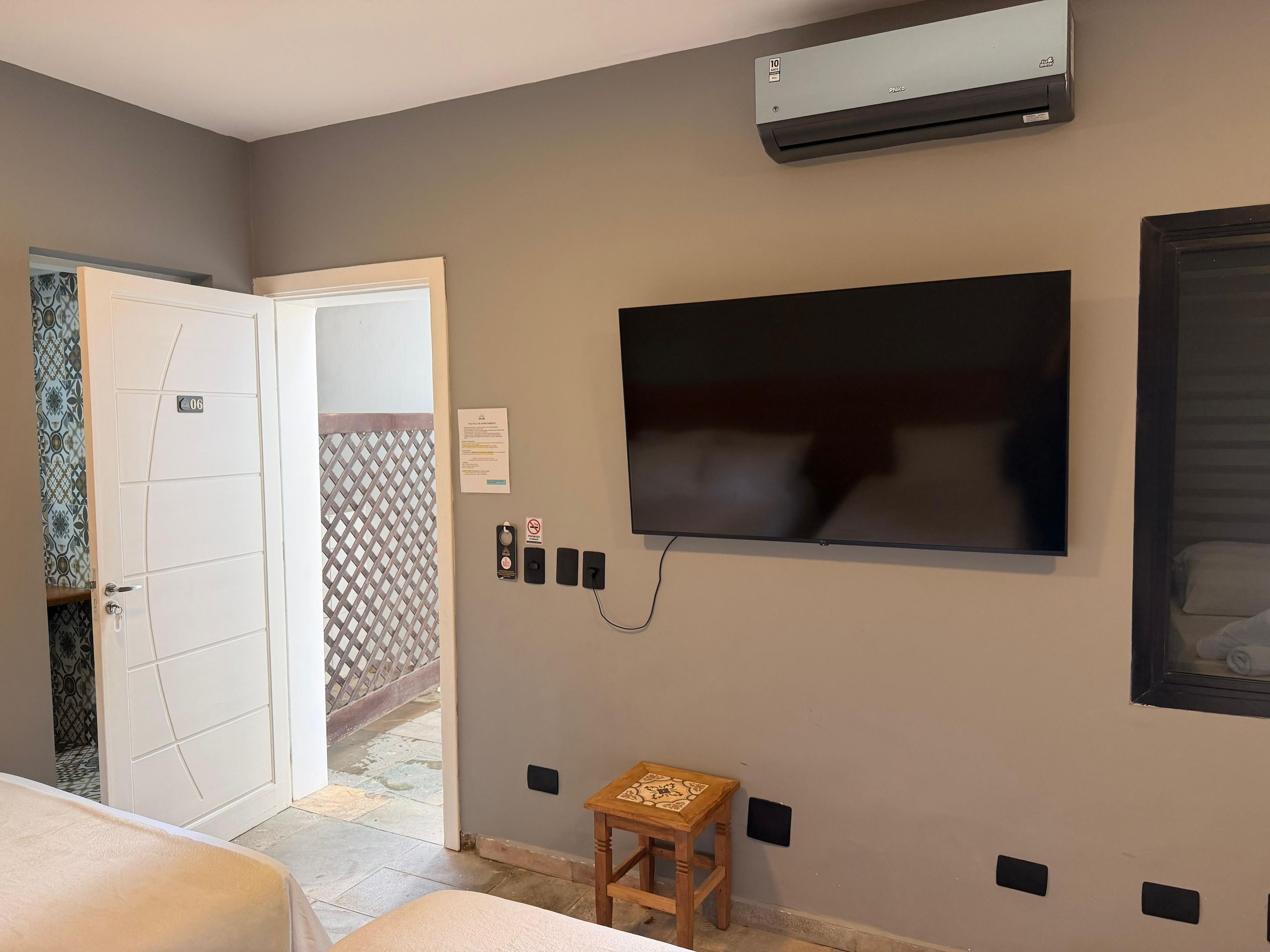 Ein Hotelzimmer mit einem an der Wand befestigten Flachbildfernseher, einer Tür mit einer Nummertafel, einem kleinen Holzschemel und einem gefliesten Boden.