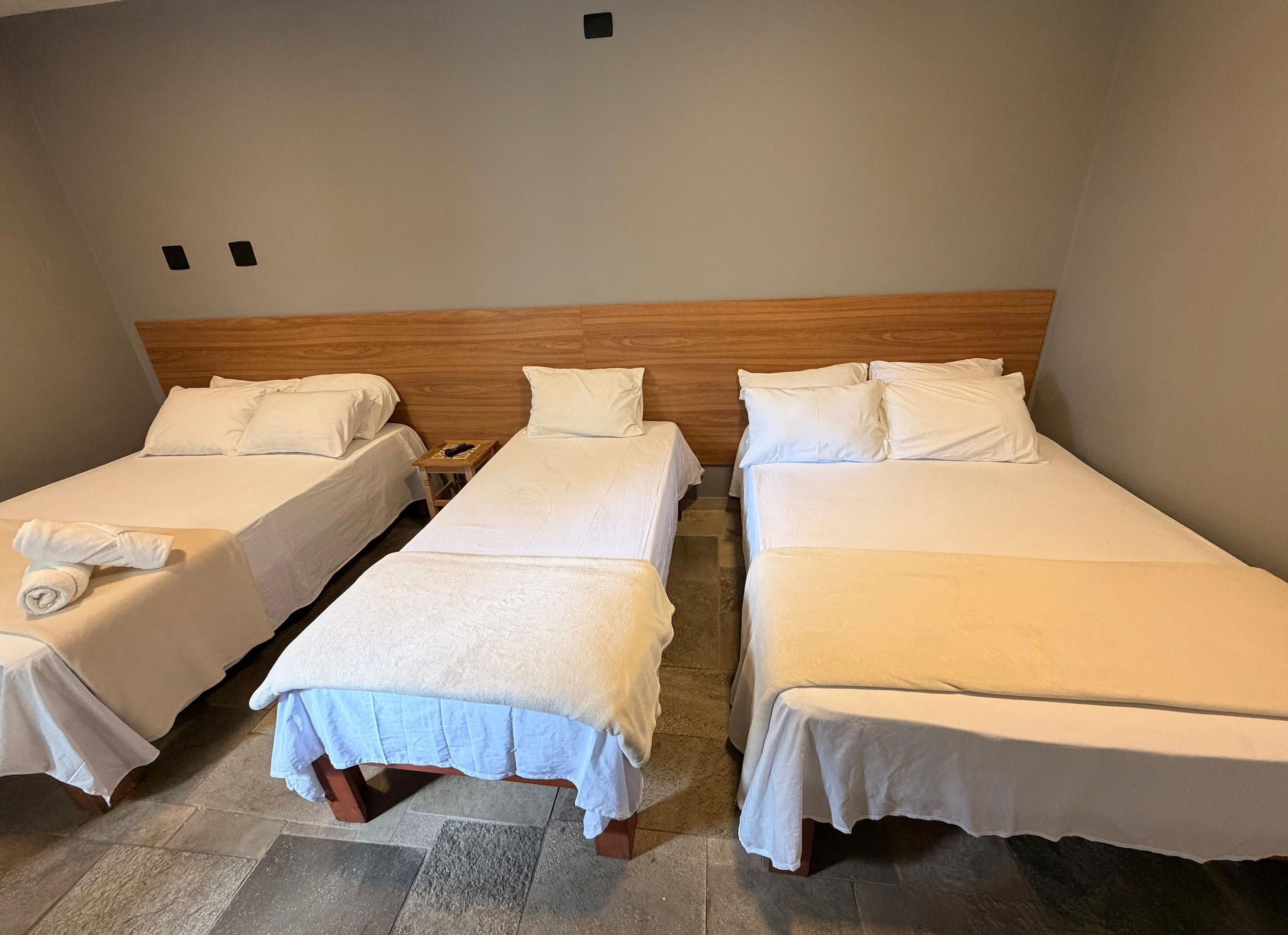 Ein Hotelzimmer mit drei Einzelbetten, jeweils mit weißer Bettwäsche und Kissen, einem Holz-Kopfteil und einem kleinen Nachttisch mit Lampe.