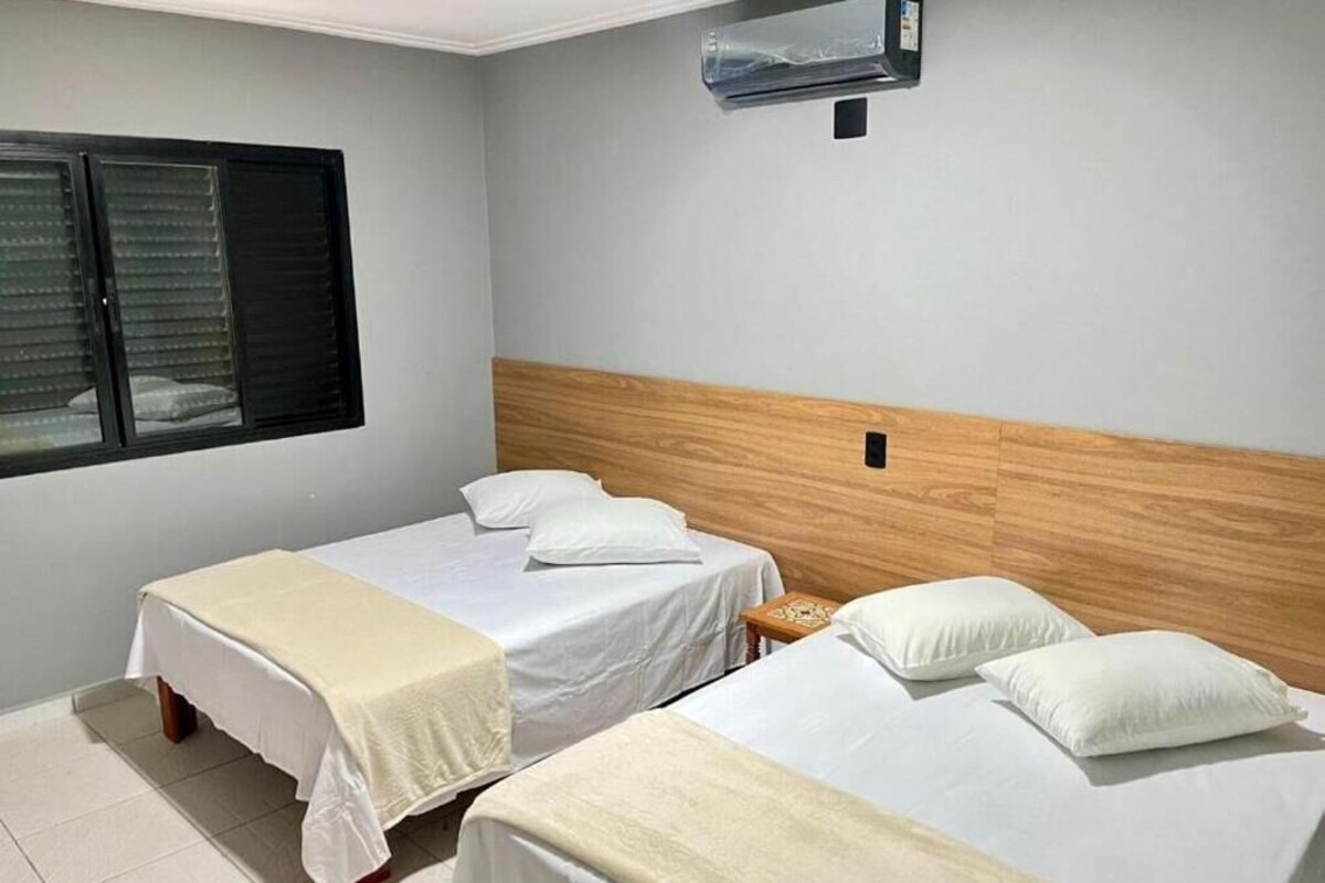 A hotel room with two single beds, a wooden headboard, a window with blinds, and an air conditioning unit.