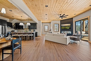 A modern kitchen with wooden ceiling and black cabinets, a dining area with a wooden table and chairs, and a living room with a sofa and a TV. - BRAND NEW Lux Cabin - Pickleball, Swim Spa + Views (Sevierville)