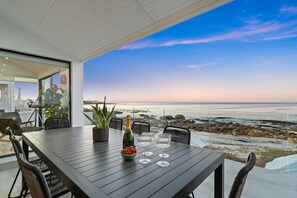 Ein moderner Essbereich im Freien mit Blick auf den Ozean, ein Tisch mit Stühlen, eine Champagnerflasche und eine Obstschale.