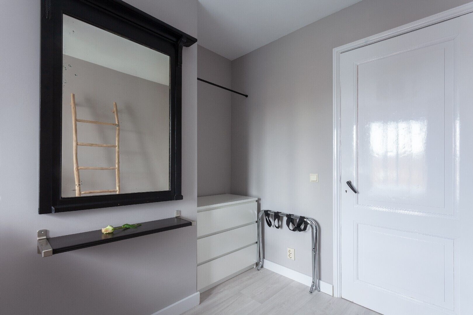 Een kamer met een grote spiegel, een witte deur, een metalen rek en een klein plankje met een plantje.