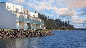 Ein Wohngebäude am Wasser mit mehreren Balkonen, einer Stein-Terrassenmauer und im Hintergrund Bäume sowie ein klarer Himmel.