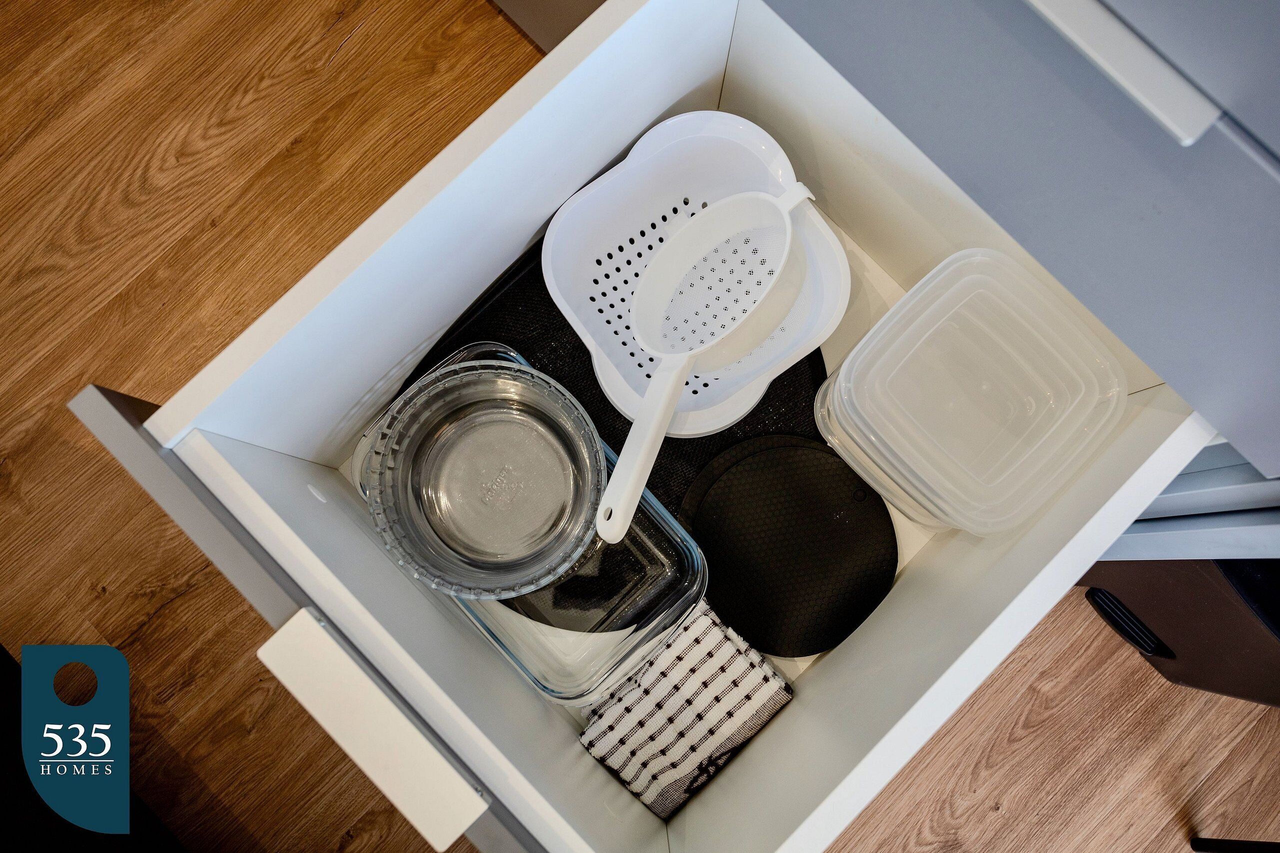 A kitchen drawer containing a colander, a plastic container, a glass jar, and a cloth.