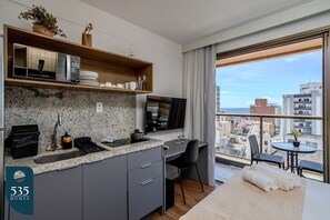 A modern hotel room with a kitchenette, a large window with a city view, a bed, and a small balcony area. - Wake up in Studio in Barra near the mall (Salvador)