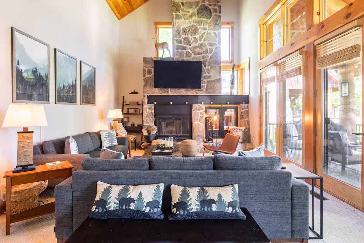 A cozy living room with a stone fireplace, a large flat-screen TV mounted above it, and a comfortable seating area with a grey sofa and armchairs.