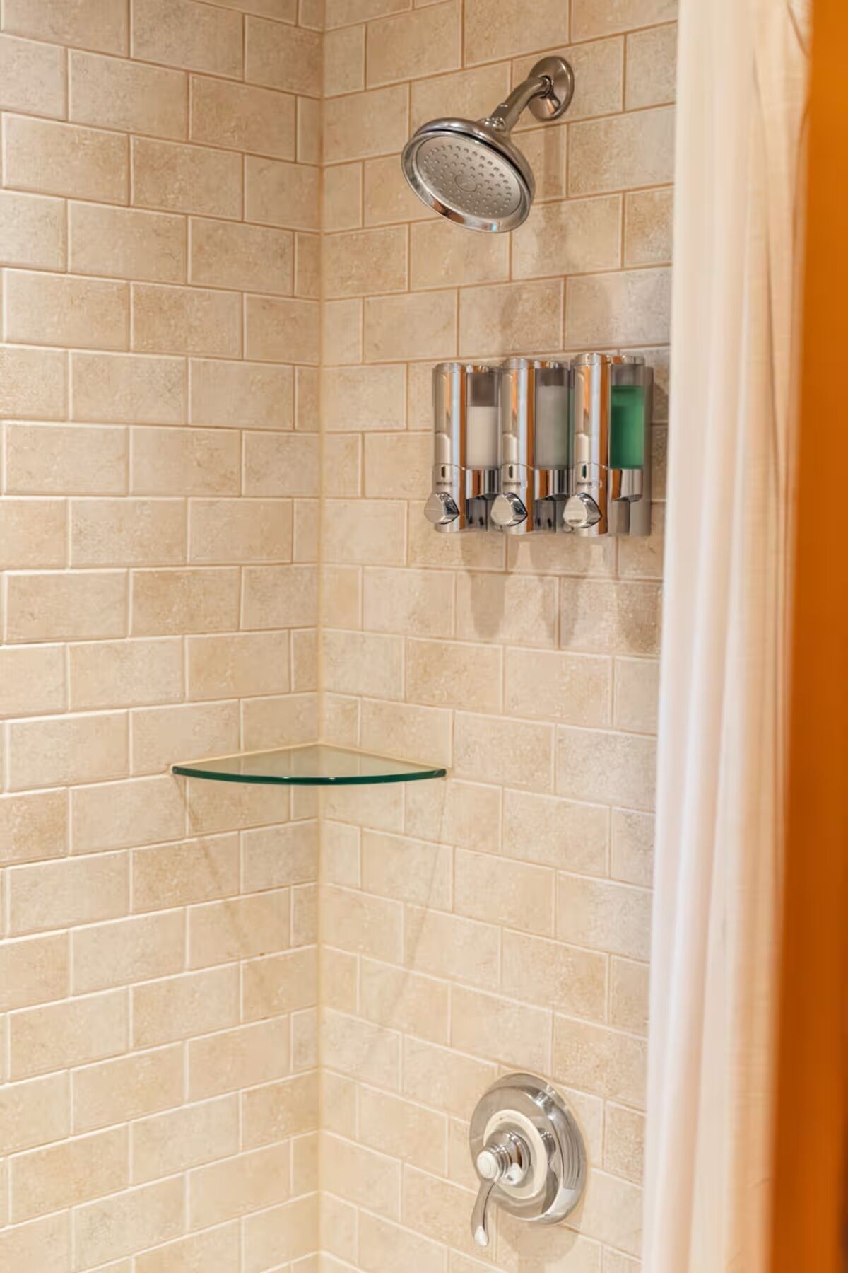 A shower area with a glass shelf, a handheld showerhead, and a wall-mounted soap dispenser.