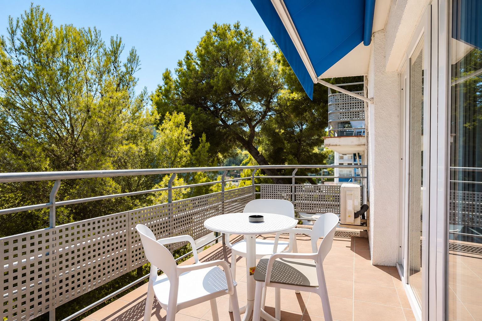A balcony with a table and chairs, a blue awning, and a view of trees and buildings.