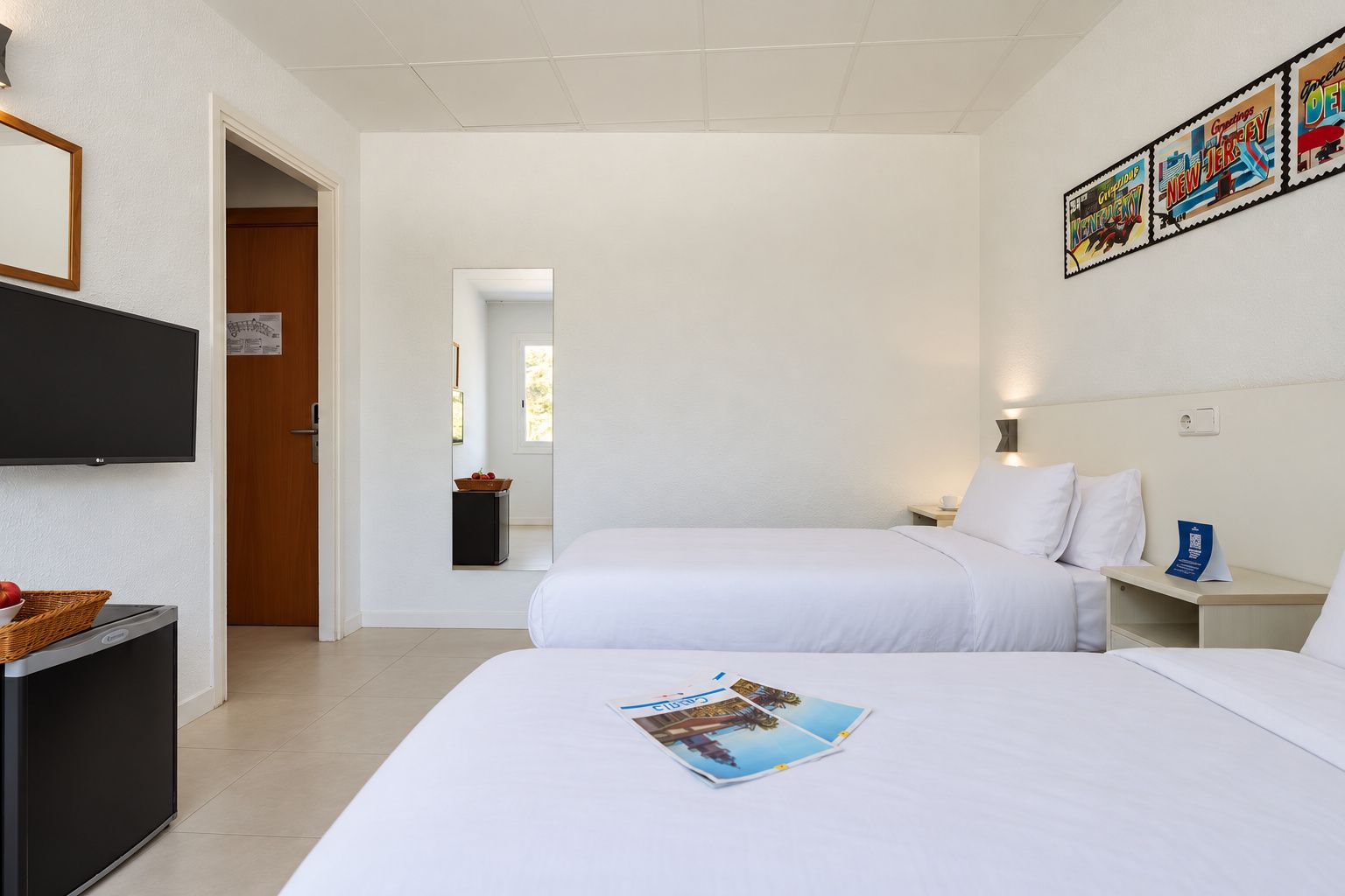 A hotel room with two beds, a television, a small table, and a wall with framed artwork.