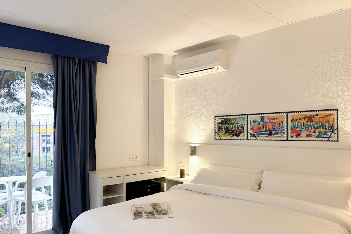 A hotel room with a bed, a desk, a chair, a window with curtains, an air conditioning unit, and a wall with three postcards.