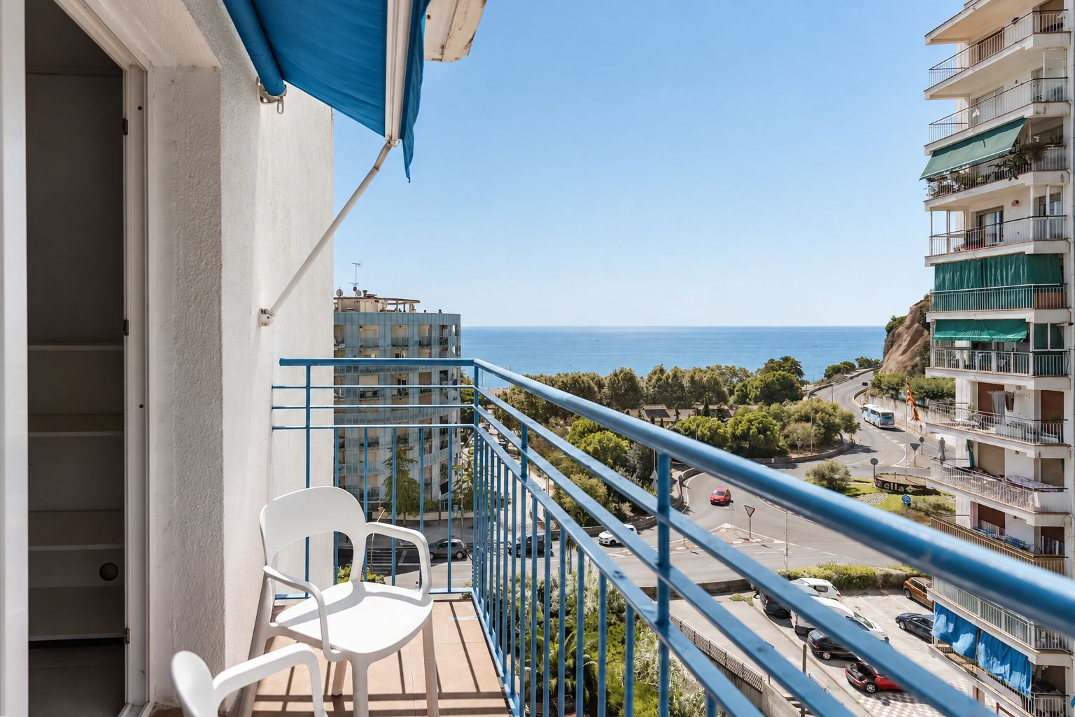 A balcony with a white chair, a blue railing, and a view of the sea and buildings.
