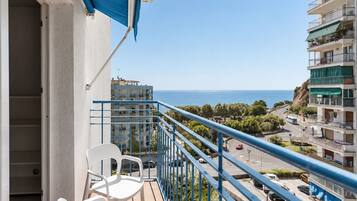 A balcony with a white chair, a blue railing, and a view of the sea and buildings.