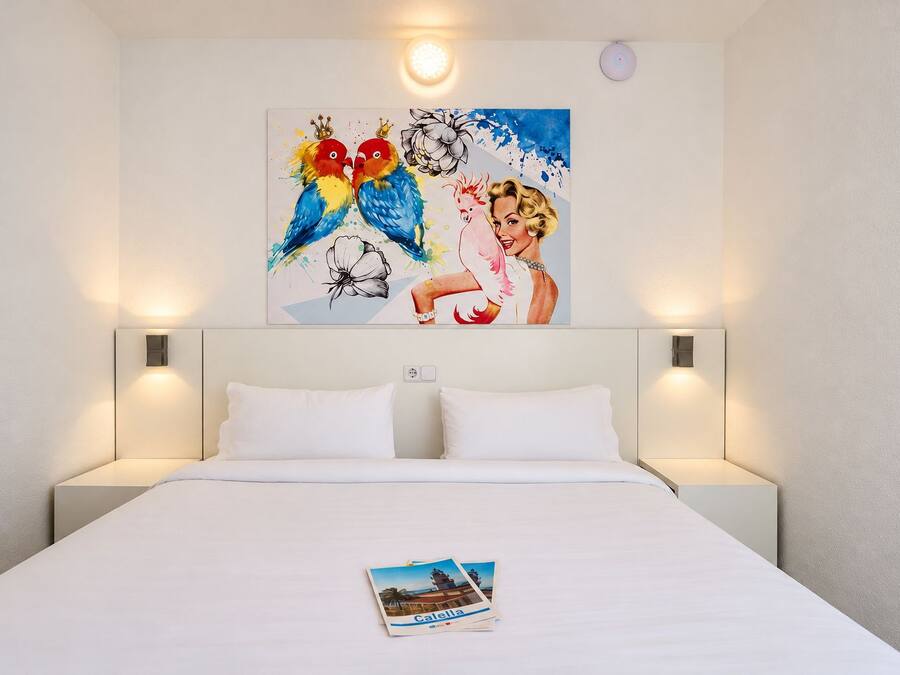 Hotellihuone, jossa on suuri sänky, kaksi yöpöytävalaisinta ja taulu seinällä.