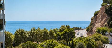 A coastal view with a road curving around a cliff, a building with balconies, and dense greenery.