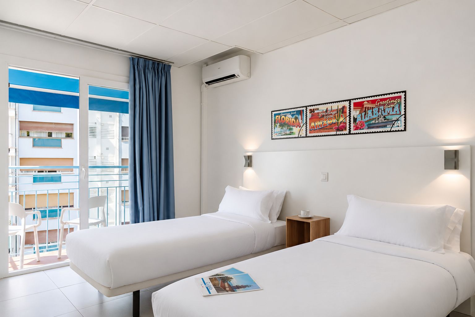 Ein Hotelzimmer mit zwei Betten, einem Fenster mit blauen Vorhängen, einer Klimaanlage und einer Wand mit drei gerahmten Postkarten.