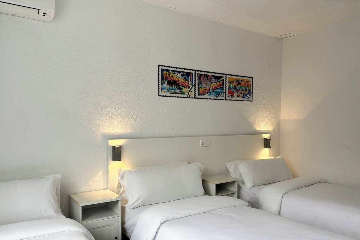 A hotel room with two single beds, a nightstand with a lamp, and a wall-mounted air conditioning unit.