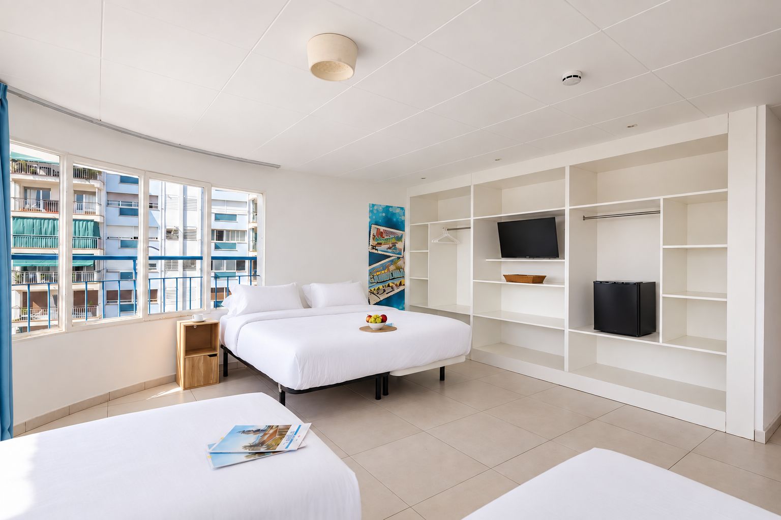 Une chambre d’hôtel équipée d’un lit, d’une télévision, d’une armoire et d’une fenêtre donnant sur un immeuble.