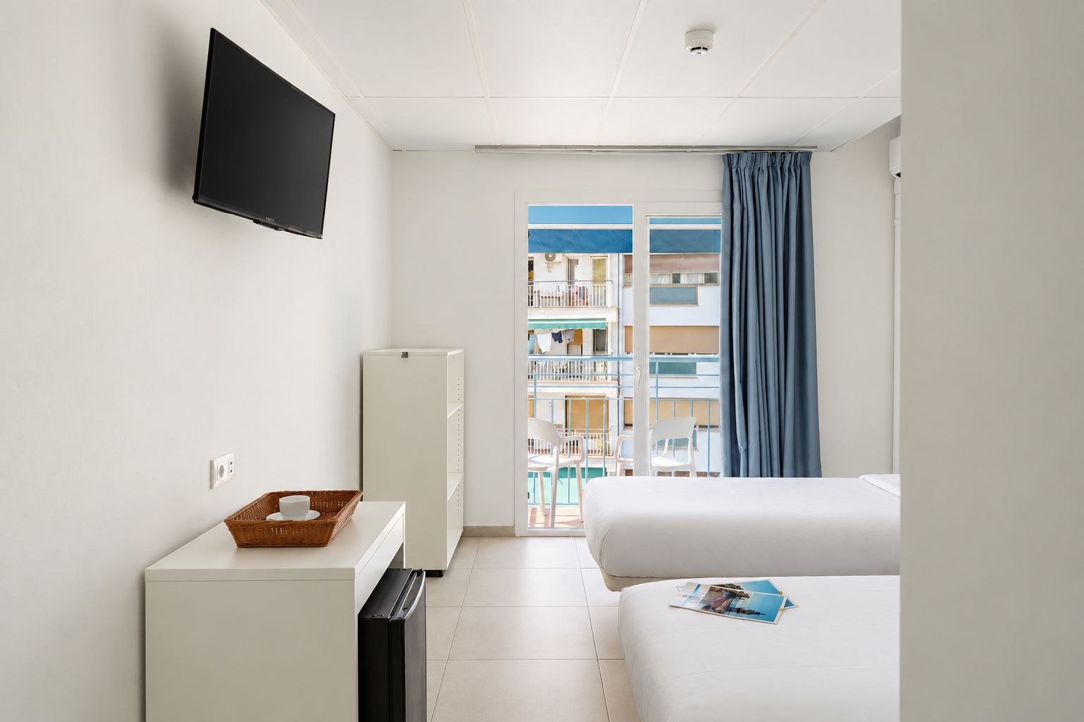Habitación de hotel con televisión, mini refrigerador, una mesita con una cesta, cama y vista a un balcón con sillas.