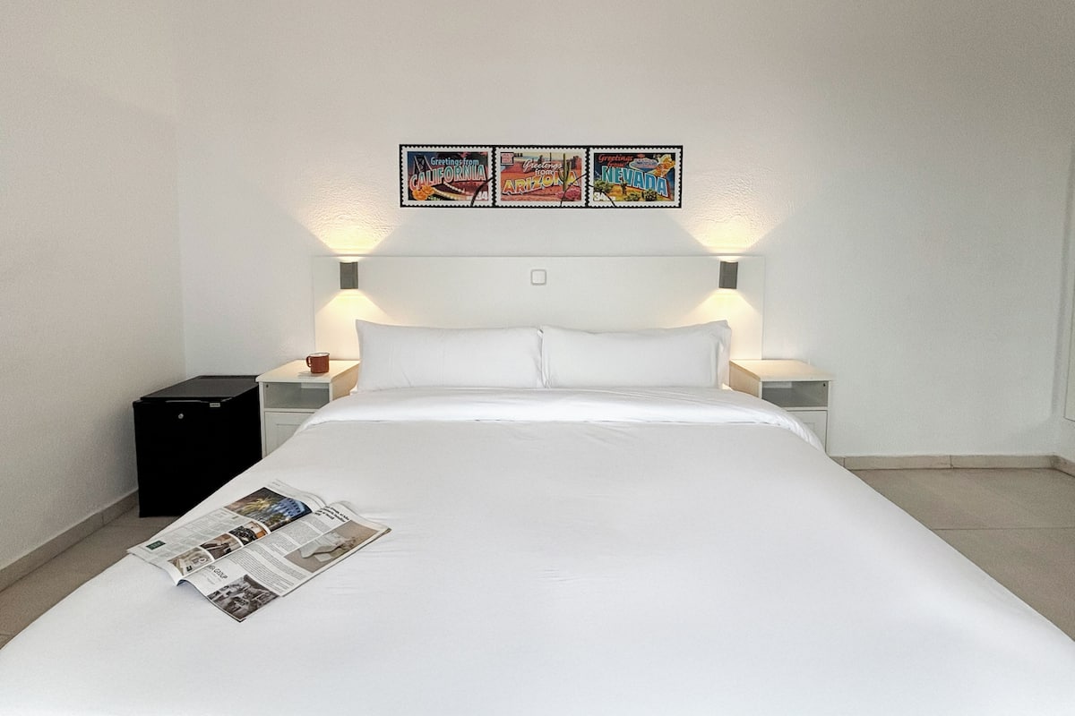 A hotel room with a bed, two bedside tables, a lamp, a small black cabinet, and three framed pictures on the wall.