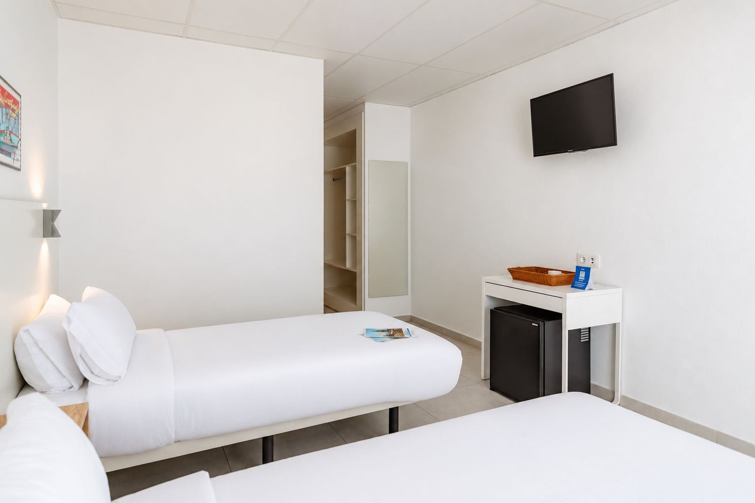 A hotel room with two beds, a small bedside table with a basket, a TV mounted on the wall, and a closet with a mirror.