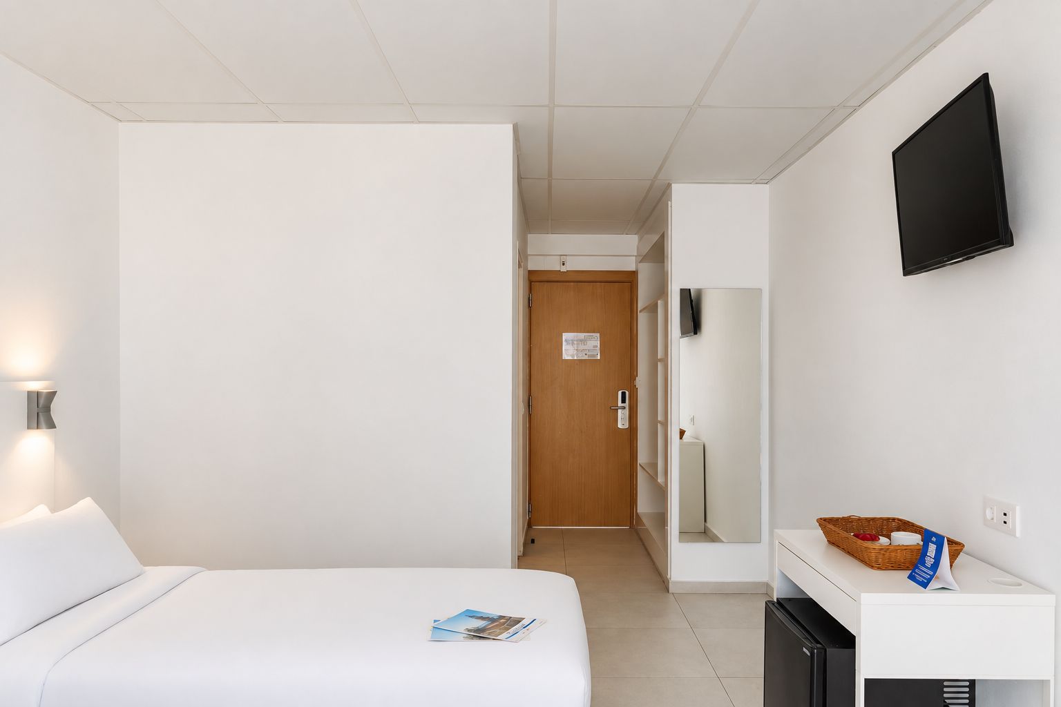 Una habitación de hotel con cama, escritorio, televisor y puerta que da a un pasillo.