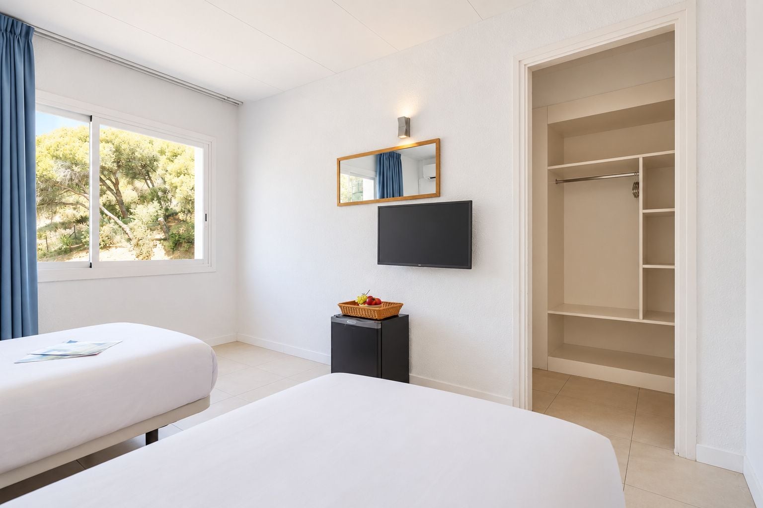 A hotel room with two beds, a television, a mirror, a small table with a basket, and a wardrobe.