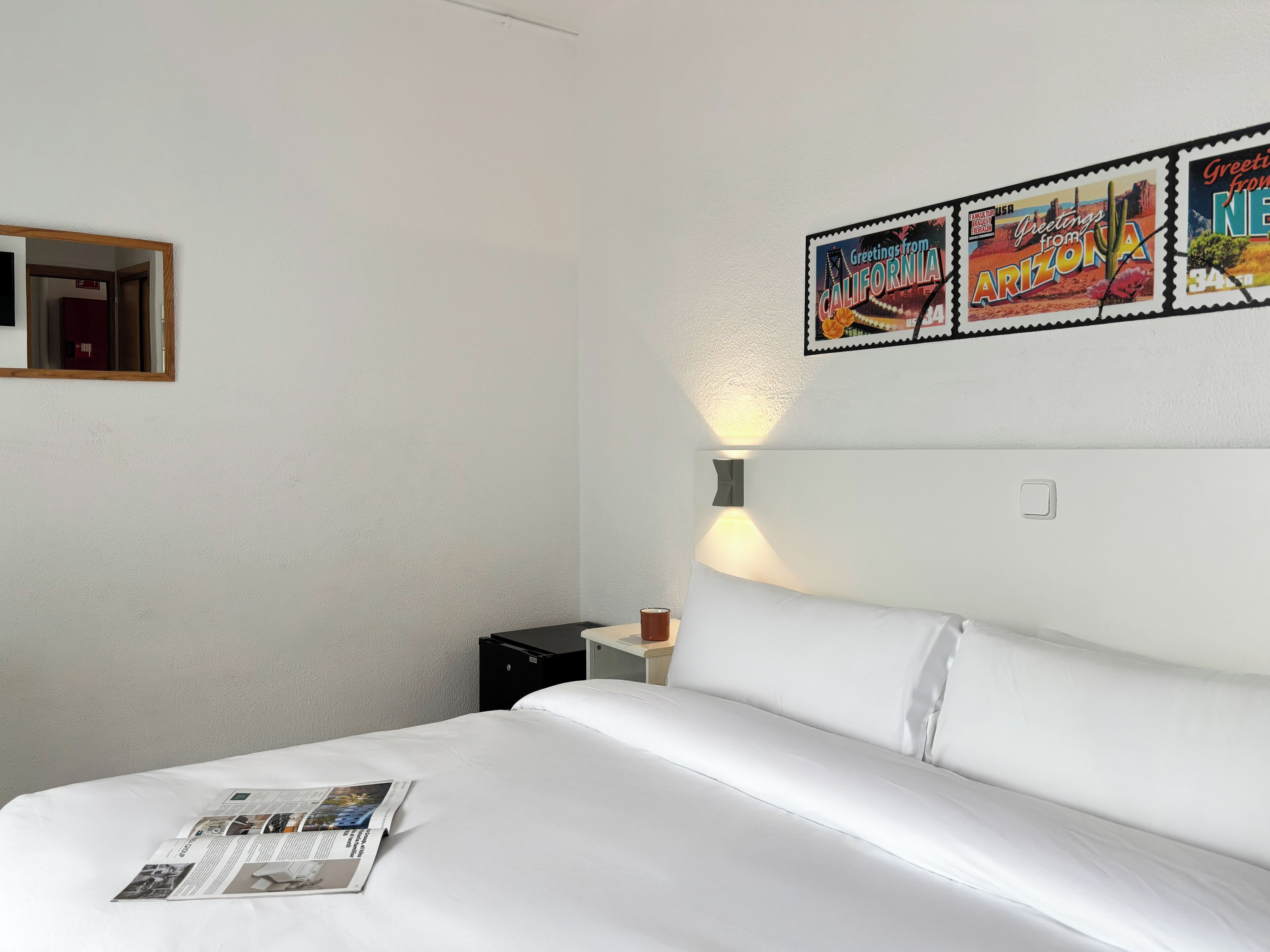 A hotel room with a neatly made bed, a wall-mounted light, and a framed picture of Arizona.