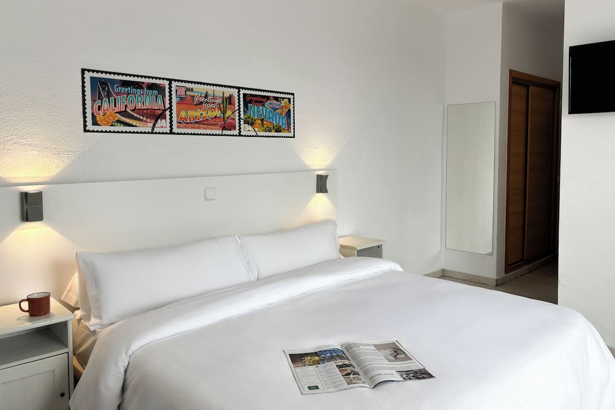 A hotel room with a bed, a nightstand, a reading light, and a wall-mounted picture of three postcards.