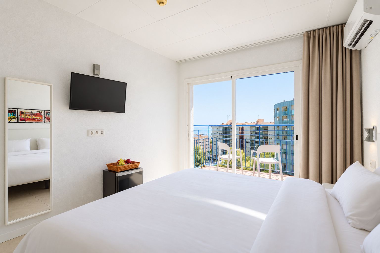 Habitación de hotel con una cama grande, un televisor en la pared, una mesita con una cesta de frutas y un balcón con sillas.