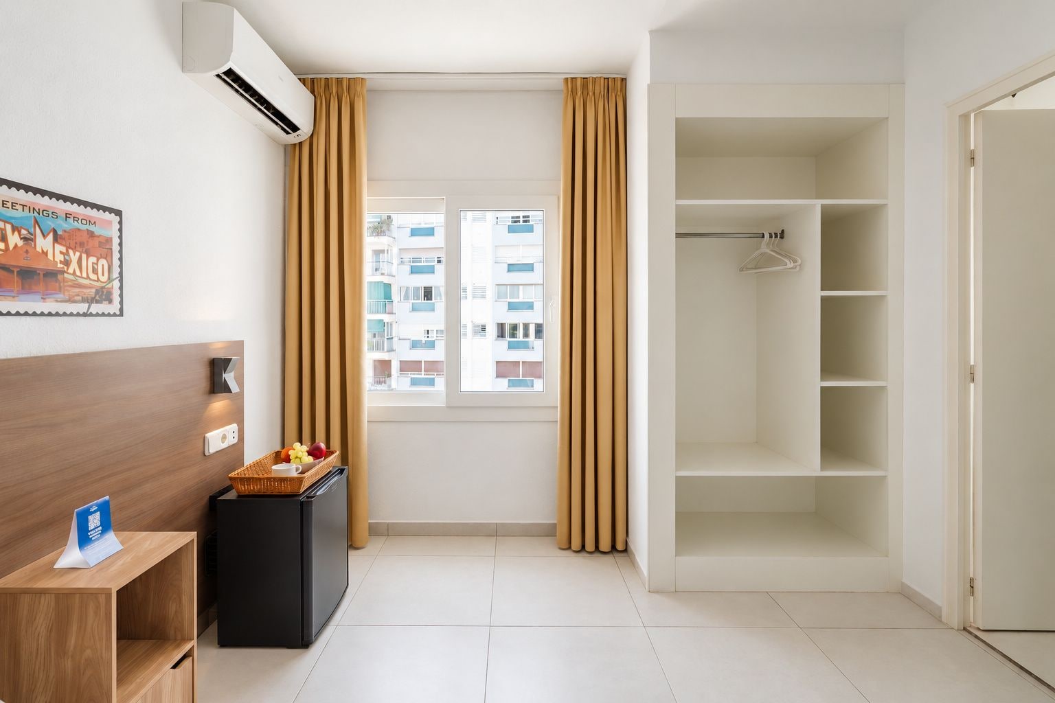 Habitación de hotel con ventana, armario, una pequeña mesa con cesta de fruta y aire acondicionado de pared.