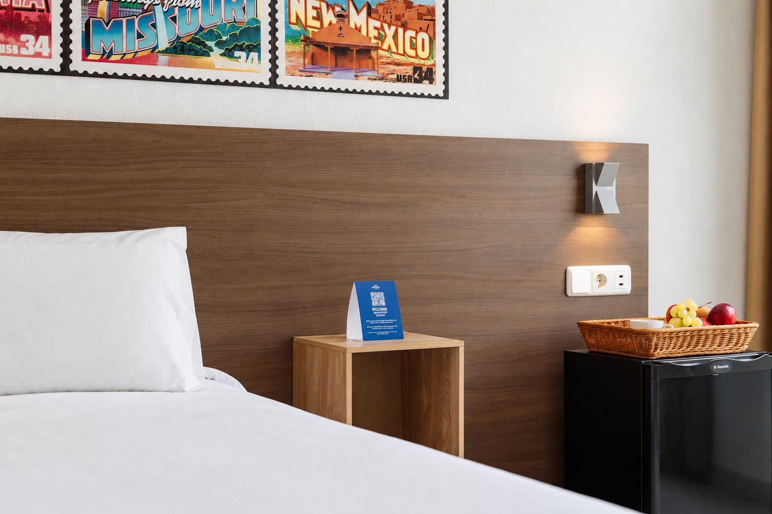 A hotel room with a wooden headboard, a neatly made bed, a small wooden nightstand with a welcome card, a basket of fruit, and a black minibar.