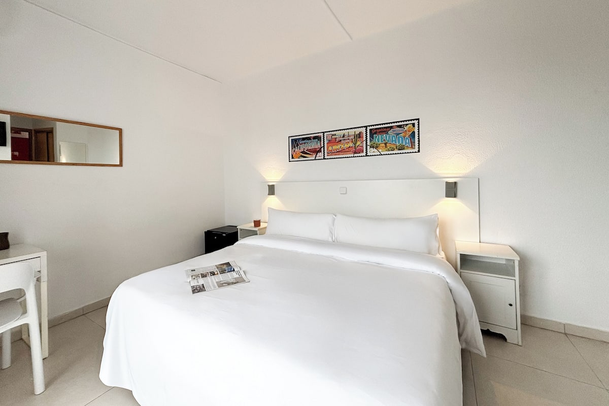 A hotel room with a bed, a desk, a chair, a nightstand, and a wall-mounted mirror.