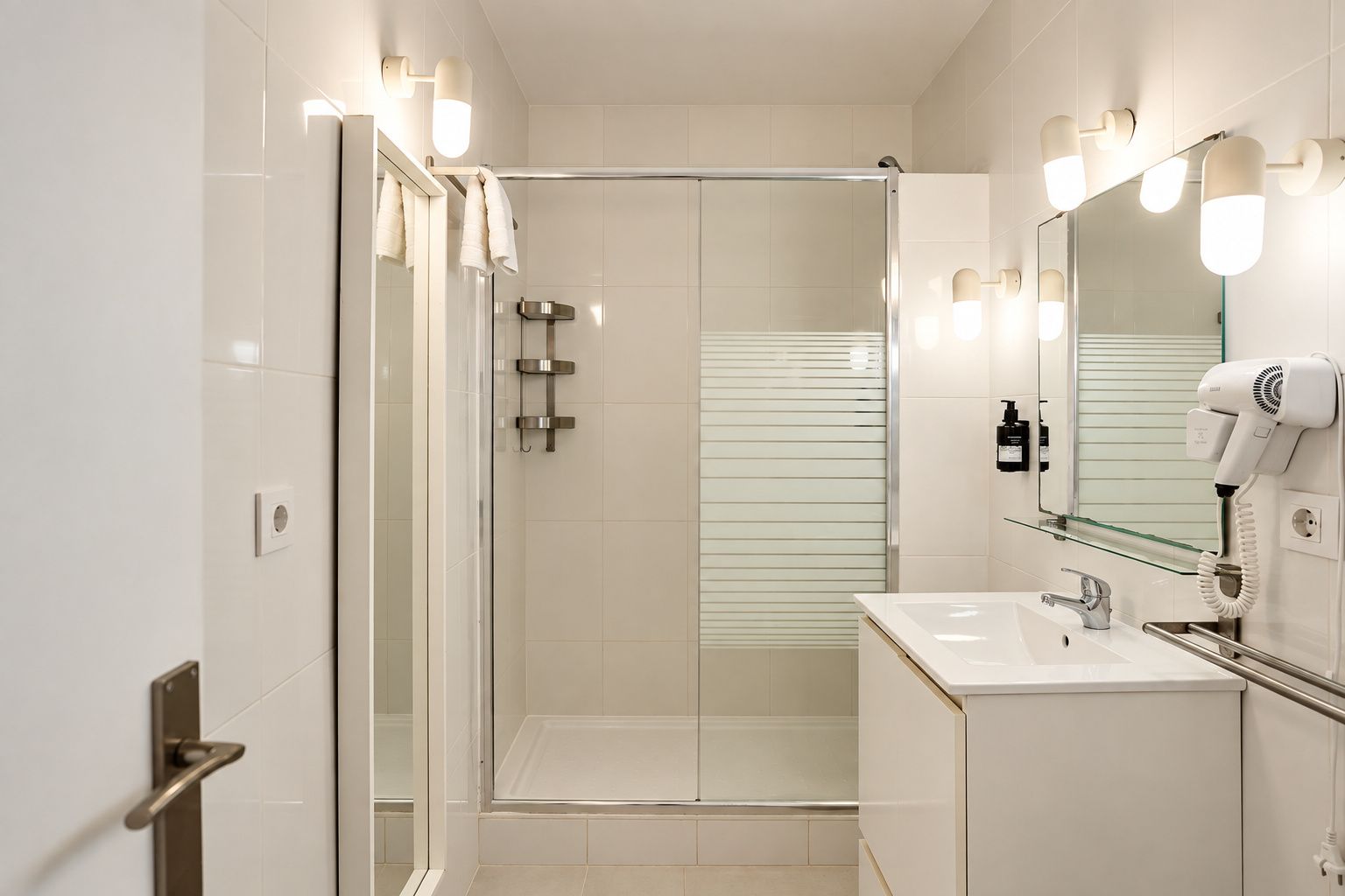 A modern bathroom with a glass-enclosed shower, a vanity with a sink, and a hairdryer mounted on the wall.