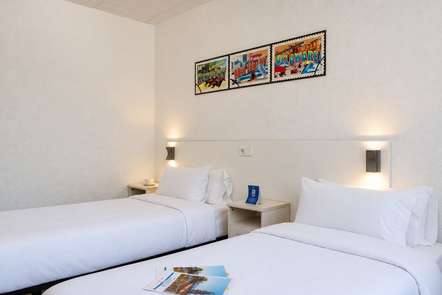 A hotel room with two single beds, a nightstand with a cup, and a wall-mounted light fixture.